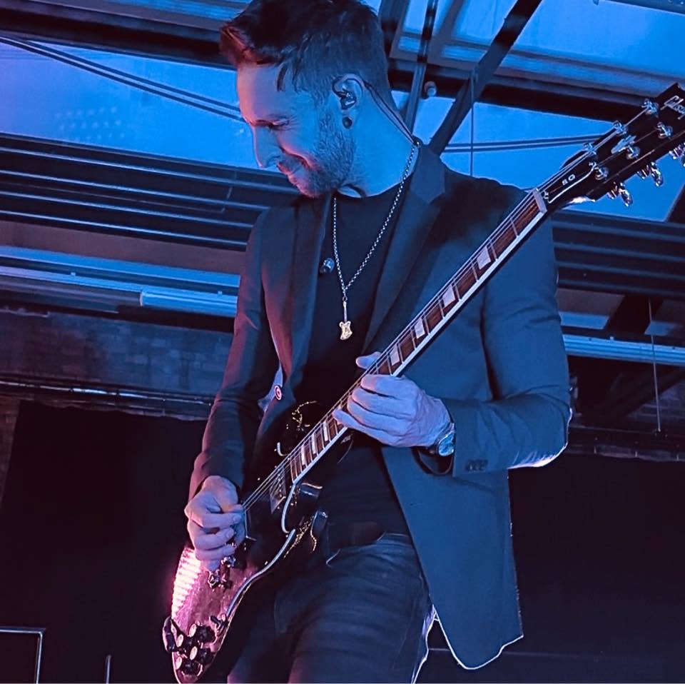 Rob Galley smiling as he plays a black electric guitar on stage under blue light