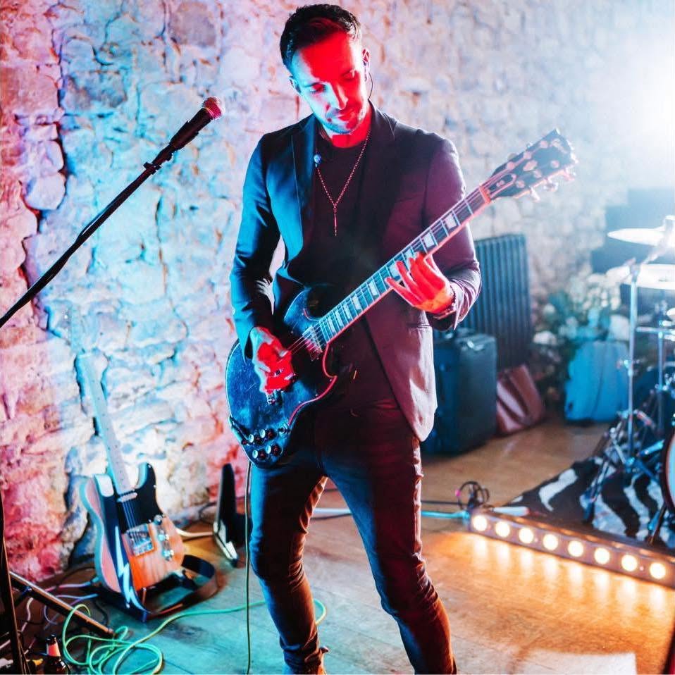 Rob Galley playing an electric guitar at a gig, in front of a stone wall lit red and blue
